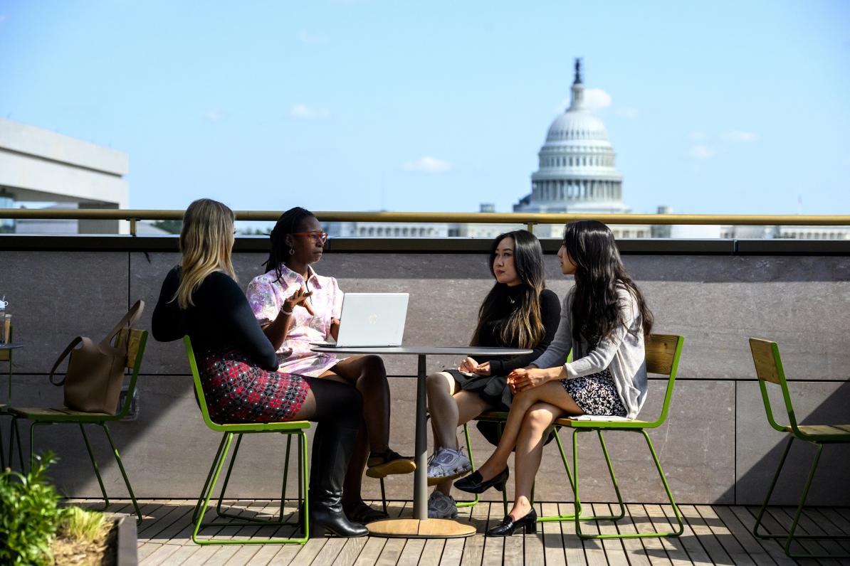 Four people at a table on the Bloomberg Center rooftop terrace, with the United States Capitol visible behind them.